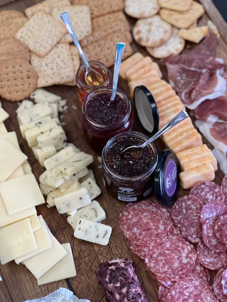 Open jars of dark fig jam on a wooden board with cheese, crackers and cured meat.