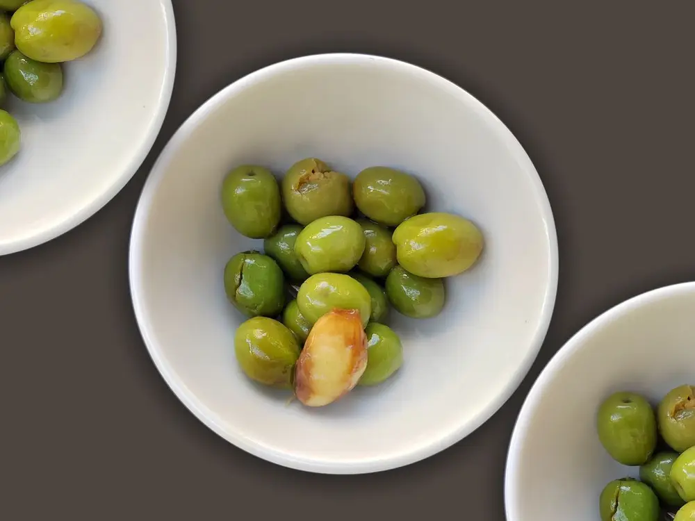A white bowl of plump green olives on a dark surface.