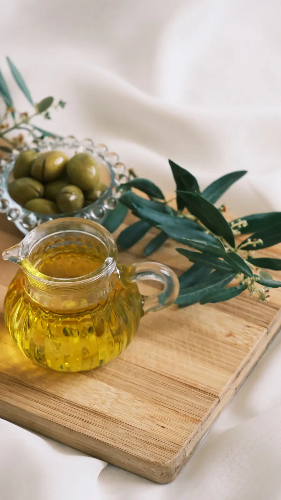 A glass jug of fresh green olive oil with olives and an olive branch on a light surface.