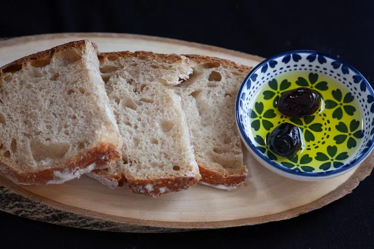 Slices of rustic bread on a wooden board beside a patterned bowl of golden olive oil with two olives.