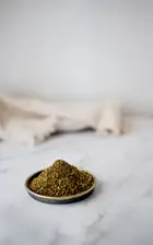 A dark bowl of green za'atar blend with sesame seeds on a marble surface beside linen.