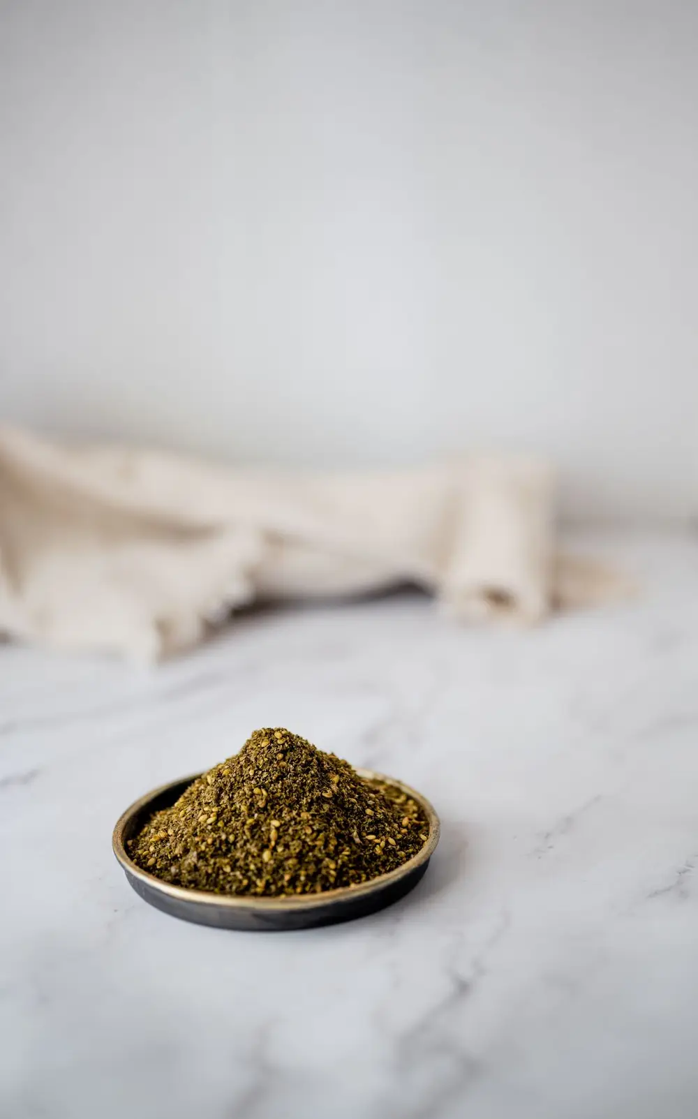 A dark bowl of green za'atar blend with sesame seeds on a marble surface beside linen.